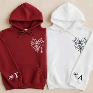 Pode incluir: Duas camisolas com capuz, uma bordô e outra branca. Cada uma apresenta um desenho de teia de aranha com um coração no centro e uma aranha. A camisola bordô tem a letra "T" na manga, e a branca tem a letra "A".