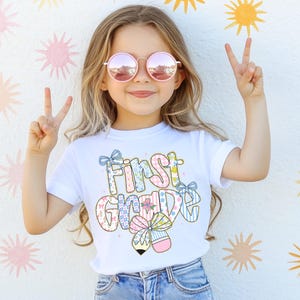 May include: A young girl wearing pink sunglasses and a white t-shirt with the words "First Grade" in a colorful, patterned design. The girl is smiling and making a peace sign with her fingers.