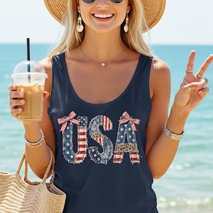 Könnte beinhalten: Ein ärmelloses, marineblaues Tanktop mit dem Schriftzug USA in einem Muster aus verwaschenem Denim, amerikanischer Flagge und Leopardenmuster. Die Buchstaben sind oben am U und A mit rot-weiß gestreiften Schleifen verziert, was ein patriotisches Design ergibt.