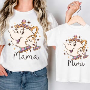 May include: White t-shirts featuring a cartoon teapot design with a smiling face and the words "Mama" and "Mimi" in black script. The teapot has gold, pink, and blue accents. The shirts are displayed on a hanger.