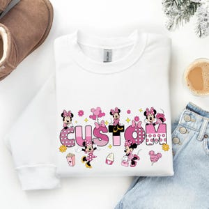 A white long-sleeved shirt with a custom text design featuring Minnie Mouse characters, a pair of jeans, and a pair of boots, along with a cup of coffee and a pair of snow boots.