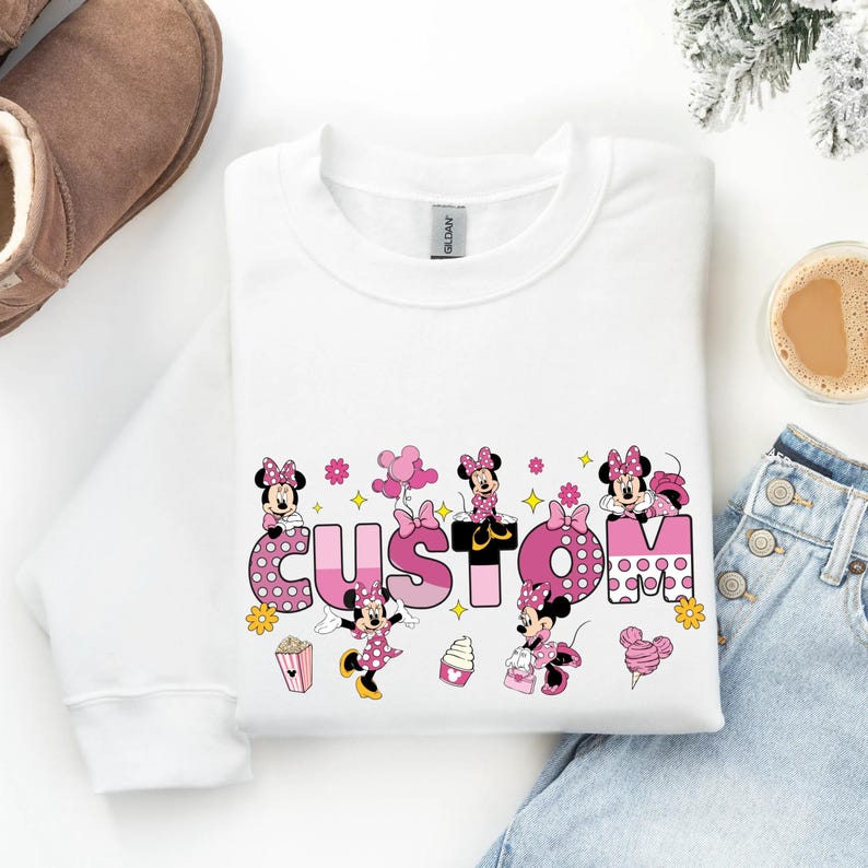 A white long-sleeved shirt with a custom text design featuring Minnie Mouse characters, a pair of jeans, and a pair of boots, along with a cup of coffee and a pair of snow boots.