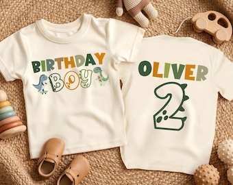 Könnte beinhalten: Zwei cremefarbene Kleinkinder-T-Shirts. Ein Shirt trägt die Aufschrift "BIRTHDAY BOY" mit Dinosaurier-Grafiken. Das andere Shirt trägt die Aufschrift "OLIVER" und die Zahl "2". Die Shirts liegen auf einer gewebten Oberfläche mit Spielzeugzubehör.