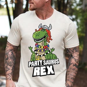 May include: A white t-shirt with a green dinosaur wearing a viking hat and a peace sign necklace. The text "Partysaurus Rex" is printed on the shirt.