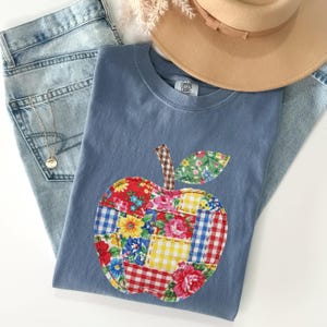 May include: A blue t-shirt featuring a large apple graphic made of various colorful fabric patches including floral, gingham, and plaid patterns. The shirt is layered over light blue denim jeans and sits beneath a tan wide-brimmed hat.