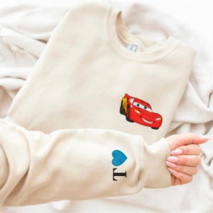 A person is holding a beige sweater with a red car embroidered on it, and there is a blue heart embroidered on the sleeve.