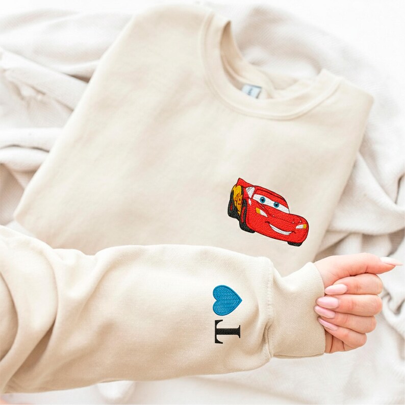 A person is holding a beige sweater with a red car embroidered on it, and there is a blue heart embroidered on the sleeve.