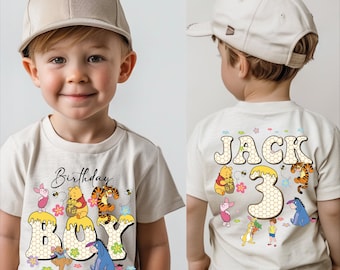 May include: A beige t-shirt with a Winnie the Pooh theme. The front features the words "Birthday Boy" with the number "3" and various characters. The back of the shirt says "Jack 3" with more characters. The boy is wearing a beige baseball cap.