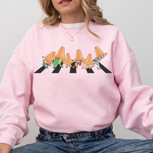 May include: Pink sweatshirt with a graphic of cartoon characters wearing traffic cones walking across a crosswalk. The characters are wearing traffic cones on their heads and are walking in a line.