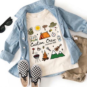 Op de afbeelding: Een crèmekleurig t-shirt met een kampeerthema, inclusief een tent, zon en kaart, wordt gecombineerd met een spijkeroverhemd, een kaki short en schoenen met een schaakbordpatroon. De tekst op het shirt luidt "Custom Crew Location 2026."