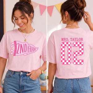 Op de afbeelding: Een lichtroze t-shirt met een afbeelding op de voorkant van een wimpel met de tekst 2ND GRADE in donkerroze. Op de achterkant staat de tekst MRS. TAYLOR boven het getal 02 in een groot, donkerroze en wit geblokt patroon.
