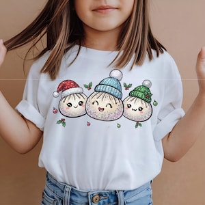 Könnte beinhalten: Ein weißes T-Shirt mit einer Grafik von drei lächelnden, glitzernden Knödel-Figuren. Der linke Knödel trägt eine rote Weihnachtsmütze, der mittlere eine hellblaue Strickmütze und der rechte eine grüne Strickmütze, alle mit Stechpalmen verziert.