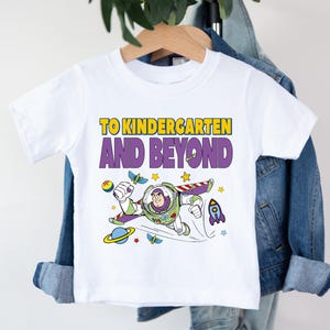 A white t-shirt with a colorful graphic of a character from the Toy Story franchise, along with the text "TO KINDERGARTEN AND BEYOND" printed on it.
