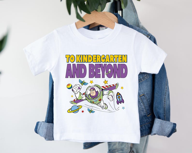 A white t-shirt with a colorful graphic of a character from the Toy Story franchise, along with the text "TO KINDERGARTEN AND BEYOND" printed on it.