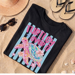 May include: Black t-shirt with a colorful graphic of a sea turtle and the text "Turtles Over It." The turtle is pink, blue, and green, with flowers and stars. The shirt is on a sandy surface with sunglasses, a straw hat, and sandals.
