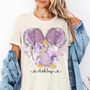 May include: A cream colored t-shirt with a purple watercolor design of a Disney princess with long blonde hair and a purple dress. The design is in the shape of a Mickey Mouse head with a large purple bow. The text "ashley" is written in cursive below the design.