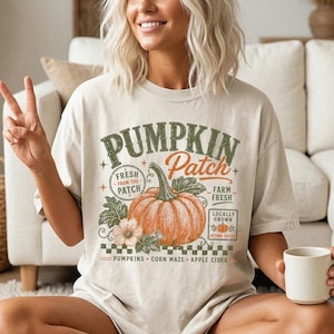 May include: A beige short-sleeve t-shirt featuring a graphic with the text PUMPKIN Patch, FRESH FROM THE PATCH, FARM FRESH, LOCALLY GROWN, and PUMPKINS CORN MAZE APPLE CIDER. The design includes a large orange pumpkin and a small flower.
