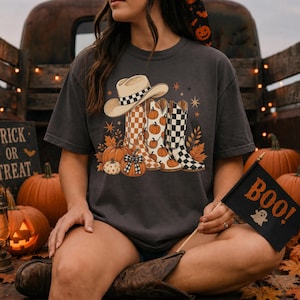 May include: A charcoal gray t-shirt featuring a graphic print of a cowboy hat and two western boots. The boots are decorated with pumpkin and checkerboard patterns. The design includes orange pumpkins, autumn leaves, and small stars.