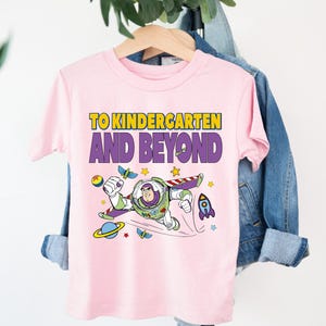 A pink t-shirt with a cartoon character and text printed on it, hanging on a wooden hanger.