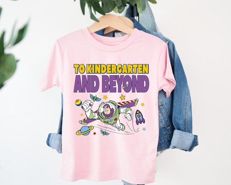 A pink t-shirt with a cartoon character and text printed on it, hanging on a wooden hanger.