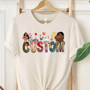 May include: A beige t-shirt with a colorful graphic design featuring characters from the animated film Moana. The graphic includes Moana, Maui, Pua the pig, Hei Hei the rooster, and the word "CUSTOM".