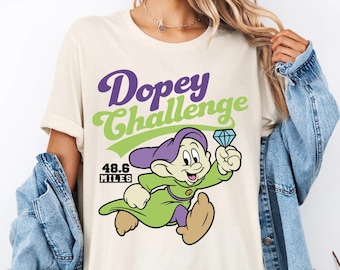 May include: Cream-colored t-shirt featuring the text "Dopey Challenge" in purple and green, with a cartoon image of Dopey holding a diamond. The shirt also displays "48.6 MILES".