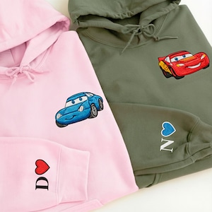 two hooded sweatshirts with cartoon car designs on them. The hooded sweatshirt on the left is pink, while the hooded sweatshirt on the right is olive green.