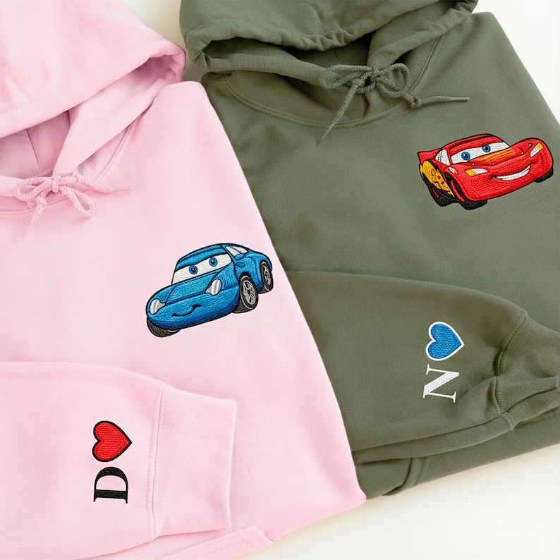 two hooded sweatshirts with cartoon car designs on them. The hooded sweatshirt on the left is pink, while the hooded sweatshirt on the right is olive green.
