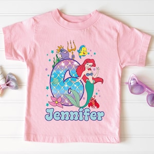 A pink t-shirt with a mermaid design and the number 6, along with a bow, sunglasses, and a unicorn bow, all arranged on a white background.