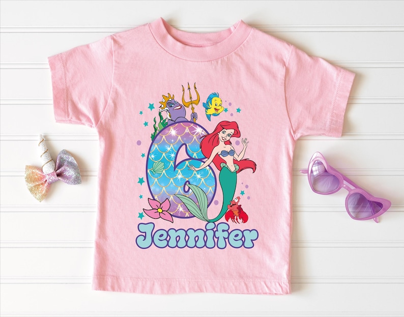 A pink t-shirt with a mermaid design and the number 6, along with a bow, sunglasses, and a unicorn bow, all arranged on a white background.