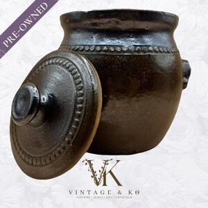 Puede incluir: Un tarro de cerámica marrón oscuro usado con tapa. El tarro tiene un cuerpo redondeado con un borde ligeramente ensanchado y dos asas pequeñas. La tapa está parcialmente abierta, revelando el interior. El tarro está decorado con un diseño de cuentas.