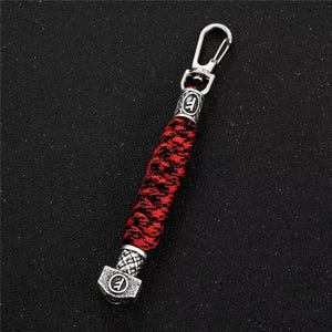 May include: A keychain with a red and black braided cord. The keychain has a silver-colored metal clasp and decorative ends. The ends feature a circular design with a symbol in the center. The keychain is approximately 5 inches long.