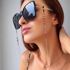 Op de afbeelding: Zwarte zonnebril met een zilveren ketting en donkergrijze kralen. De zonnebril heeft een cat-eye vorm met goudkleurige pootjes. Een zilveren ketting met een parel hanger is ook zichtbaar.