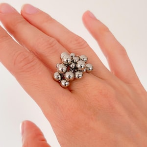 Könnte beinhalten: Ein silberfarbener Ring mit einer Ansammlung kleiner, kugelförmiger Perlen. Die Perlen sind so angeordnet, dass sie ein strukturiertes, auffälliges Design bilden. Der Ring wird an einem Finger gezeigt, um seine Größe und Form hervorzuheben.