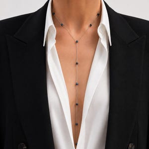 Könnte beinhalten: Eine zarte Halskette mit einer Reihe kleiner, dunkler Perlen, die an einer feinen Kette hängen. Die Halskette wird über einem weißen Hemd und einem schwarzen Blazer getragen, was einen raffinierten und eleganten Look erzeugt. Die Perlen scheinen gleichmäßig verteilt zu sein.