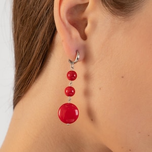 Puede incluir: Pendientes rojos colgantes con una gran cuenta redonda y cuentas redondas más pequeñas que cuelgan de un aro plateado.