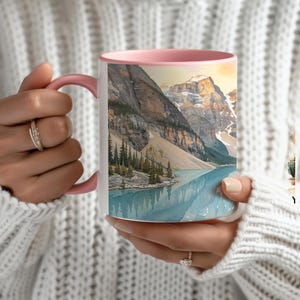 May include: A white ceramic mug with a soft pink handle and rim, featuring a detailed illustration of a mountain range with rocky peaks, evergreen trees, and a calm blue lake reflecting the landscape under a warm, golden-toned sky.