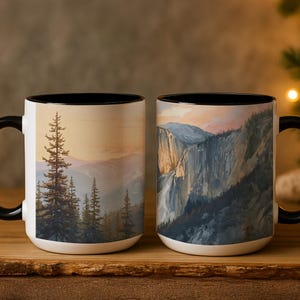 May include: Two white ceramic mugs with black handles and rims. Each mug features a landscape painting of a mountain range and trees. The artwork is in shades of green, brown, and blue, with a warm sunset sky.