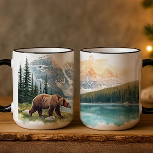 May include: Two white ceramic mugs with black handles and rims. Each mug features a watercolor painting of a mountain landscape with a lake and trees. One mug shows a brown bear. The mugs are on a wooden surface.