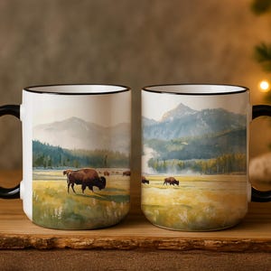 May include: Two white ceramic mugs with black handles and rims. Each mug features a watercolor painting of bison grazing in a grassy field with mountains in the background. The mugs are set on a wooden surface.