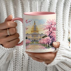 May include: A white ceramic mug with a pink handle and interior rim, featuring a watercolor-style illustration of the United States Capitol building surrounded by blooming pink cherry blossom trees under a soft, light-colored sky.