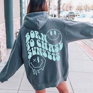 May include: A charcoal gray hoodie with a light blue graphic on the back. The graphic says "Born to Chase Sunsets" with two smiley faces.