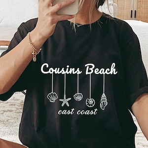 May include: Black t-shirt with the text "Cousins Beach" and "east coast" in white. Below the text are white seashell illustrations. The shirt is a casual style.