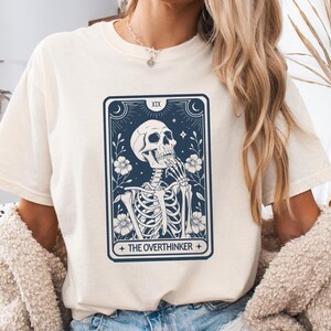May include: Ivory t-shirt with a graphic of a skeleton with the text "The Overthinker" and a tarot card design.