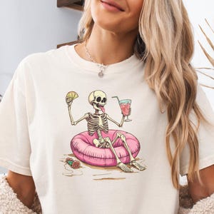 May include: Ivory-colored t-shirt featuring a skeleton relaxing on a pink inflatable ring, holding a cocktail and a lime wedge. The skeleton is in a beach setting. The shirt has the color "Ivory" printed on it.