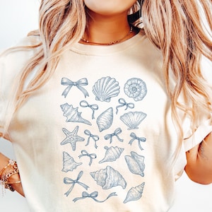 May include: Ivory-colored t-shirt featuring a collection of blue seashell illustrations and bows. The design includes various shell shapes and starfish, creating a coastal theme. The word "Ivory" is printed on a pink label.