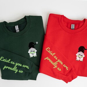 Può includere: Due felpe con un motivo a gnomo e il testo "Exist as you are, proudly so" sulla manica. Una felpa è verde bosco e l'altra è rossa.