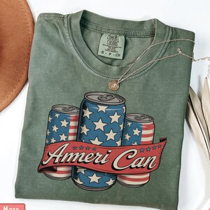 Puede incluir: Una camiseta verde musgo con un gráfico de tres latas de bebidas decoradas con estrellas y rayas. Las latas están colocadas contra una pancarta roja que dice "Ameri Can". Un collar dorado está drapeado sobre la camisa.