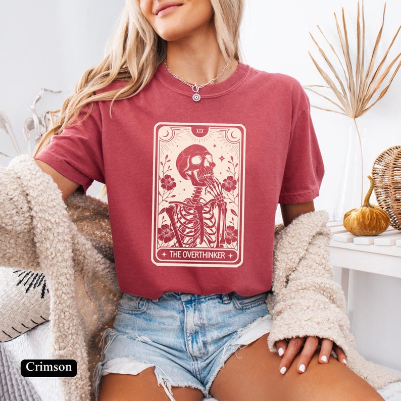 May include: A crimson t-shirt with a graphic of a tarot card featuring a skeleton with the text "The Overthinker".
