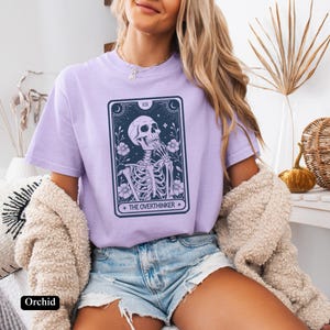 May include: A lavender t-shirt with a tarot card design featuring a skeleton with the text "The Overthinker".